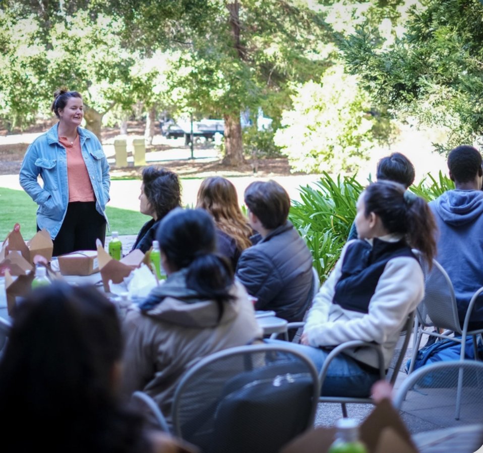 The image shows a group of people gathered outdoors in a grassy area with trees and plants around. A woman in a blue jacket and orange shirt is standing and speaking to the seated audience, who are attentively listening. There are some food items and drinks on the table in front of the group. This scene likely relates to the context of Stanford HAI student groups exploring AI safety and its impacts, as the group discussion setting aligns with the theme of collaborative exploration and dialogue on AI-related topics.